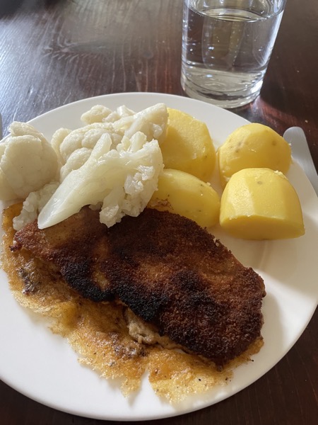 A white plate with a slightly burnt breaded pork cutlet, some cauliflower and potatoes, next to it a glass of water
