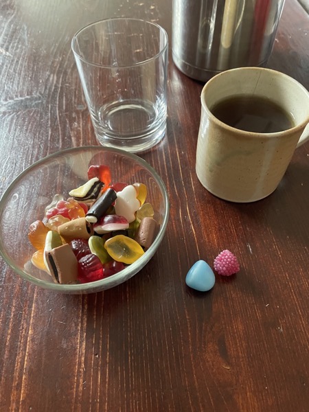 Same brown table, the glass of water is empty but there is a glass bowl holding an assortment of Haribo gummy bears and licorice with two pieces directly on the table. Next to it a mug of tea.