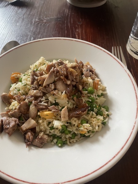 A white plate with rice salad with peas, carrots, bits of boiled egg and chicken on a brown wooden table with a glass of water on the side.