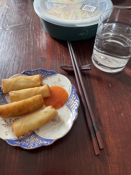 anold-fashioned blue white and gold saucer with four spring rolls and some sweet chili sauce. Next to it a pair of dark wooden chopsticks, a glass of water and a reusable container of takeout