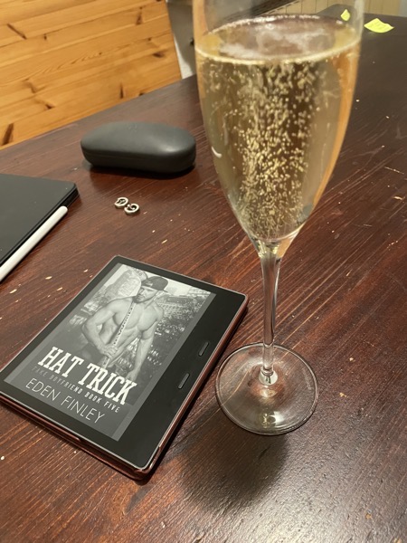 A dark brown table with an ebook reader showing the cover of "Hat Trick" by Eden Finley, a long-stemmed glass of chamapgne, some hoop earrings, a glasses case and an iPad