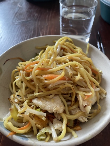 a bowl of fried noodles with chicken and veggies with a glass of water on the side and chopsticks
