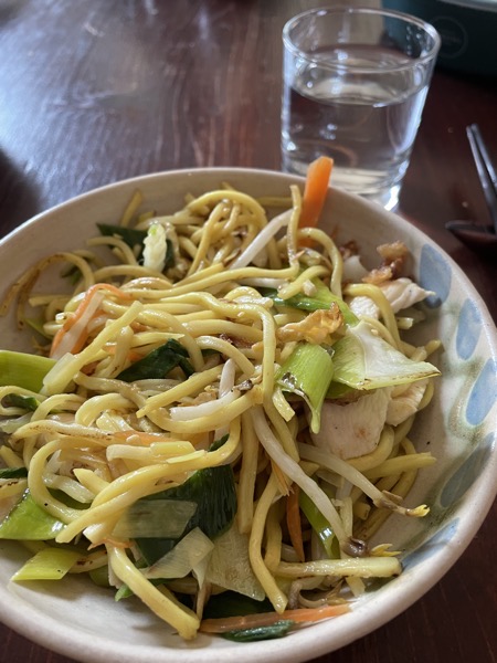 a ceramic bowl full of fried Asian noodles with veggies and chicken, dark brown chopsticks and a glass of water