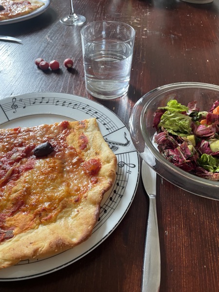 a plate with musical notes and a big slice of pizza with one olive, mixed salad, a glass of water and some radishes on a dark brown table