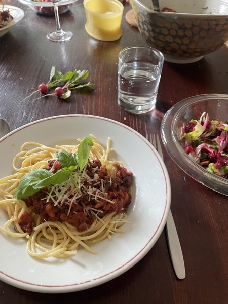 a messy table with a plate of spaghetti bolognese with prmesan and some basil leaves, a glass bowl of mixed salad, a glass of water and some turnips