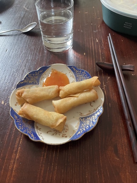 a blue, white and gold saucer with four spring rolls and some sweet chili sauce next to chopsticks and a glass of water