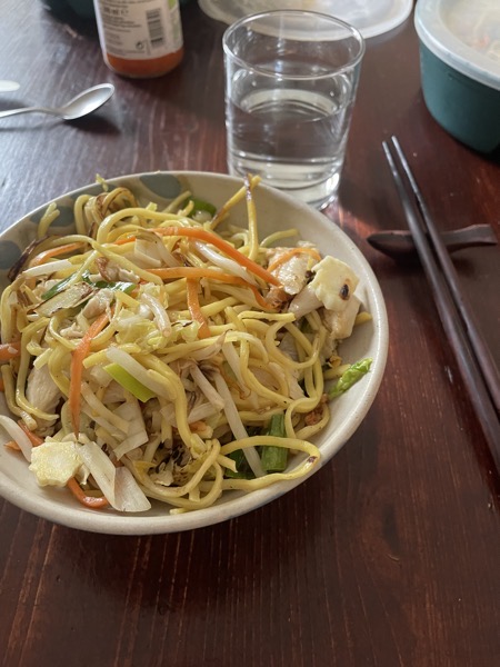 a ceramic bowl of fried noodles with veggies and chicken, same chopsticks and the same glass of water