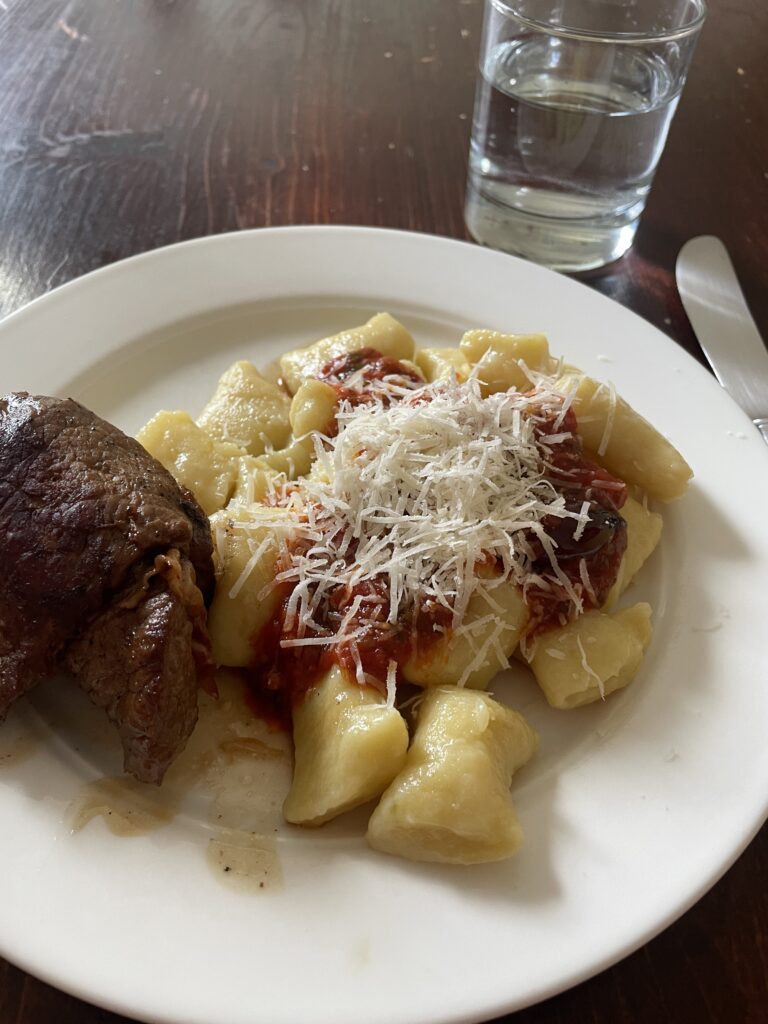 A white plate on a dark brown woode table. On the plate a rolled-up fried veal cutlet with bacon and sage next to a heap of homemade gnocchi with tomato sauce and grated parmesan. Next to it a knife and a glass of water