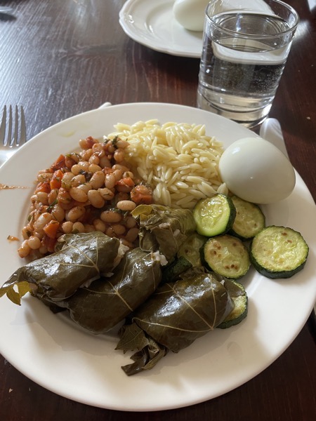 a plate with kritharaki, fried zucchini slices, a boiled egg, white beans in tomato sauce and homemade stuffed wine leaves with a glass of water