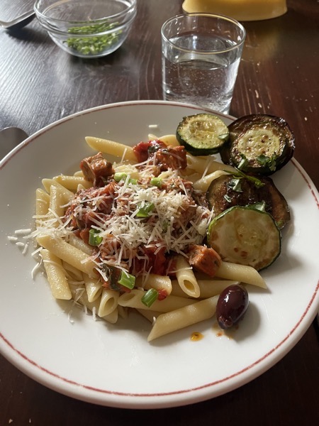 a plate of penne lisce with tomato sauce, pieces of pork cutlet, grated cheese, spring onions and fried slices of zucchini and eggplant with a glass of water