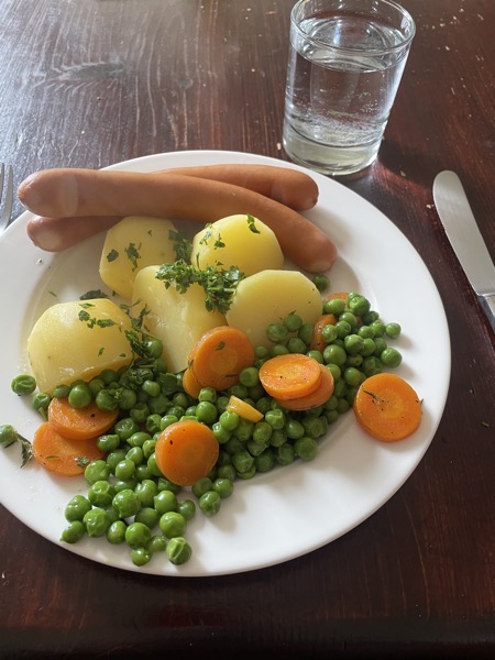 a white plate with potatoes, peas and carrots and two wieners with a glass of water