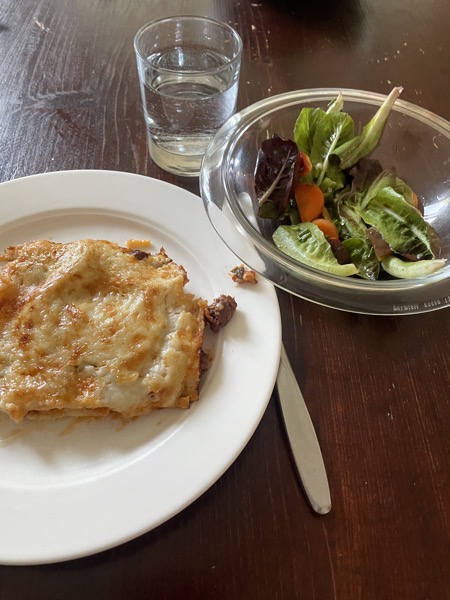 a white plate with a thin piece of lasagna, a glass bowl of salad and a glass of water on a dark brown table