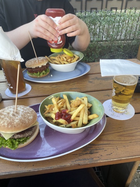 two plate of brugers and fries on a wooden table, one ice tea and one small beer, the boy is putting ketchup on his fries