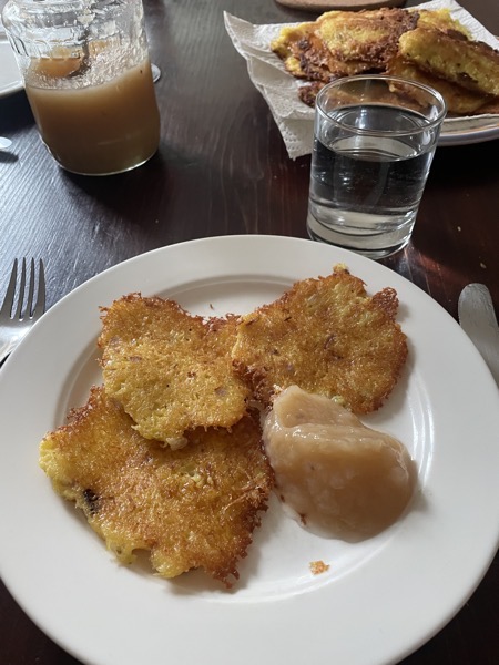 a white plate with three latkes and some applesauce on a table that also holds a glass of water, another plate with the rest of the latkes and the half-empty jar of homemade applesauce