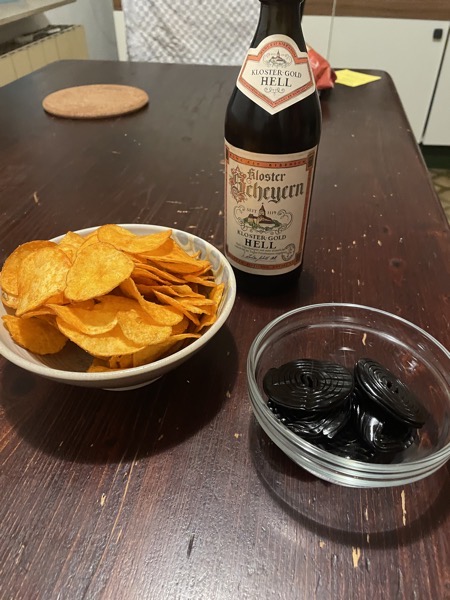 a brown wooden tabletop with a ceramic bowl full of potato chips, a smaller glass bowl with black licorice wheels and a bottle of Scheyern Hell beer