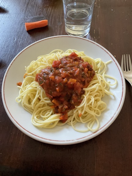 a plate of spaghetti with tomato sauce, next to it a piece of raw carrot and a glass of water