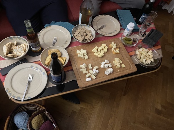 our living room table with many plates, three bowls of different kinds of crackers, a big cutting board with six kinds of cheese and a glass bowl filled with cut up pear; also diverse beverages, all of them alcohol-free for a change