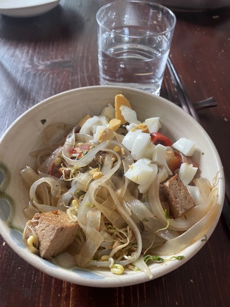 a beige ceramic bowl holding rice noodles, marinated tofu, bits of boiled egg, sprouts and all the veggies with a glass of water and a pair of chopsticks on the side