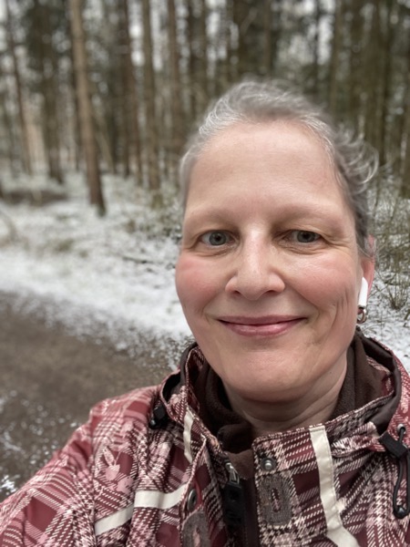 Susanne with a red face, wearing a very faded and ugly brown checkered rainjacket and no glasses in front of some pine trees sprinkled with snow