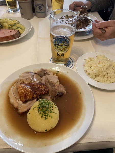 a table with a white, starched tablecloth, a plate with pork roast, sauce and a potato dumpling in the foreground paired with a half-full glass of beer and a plate of cabbage salad