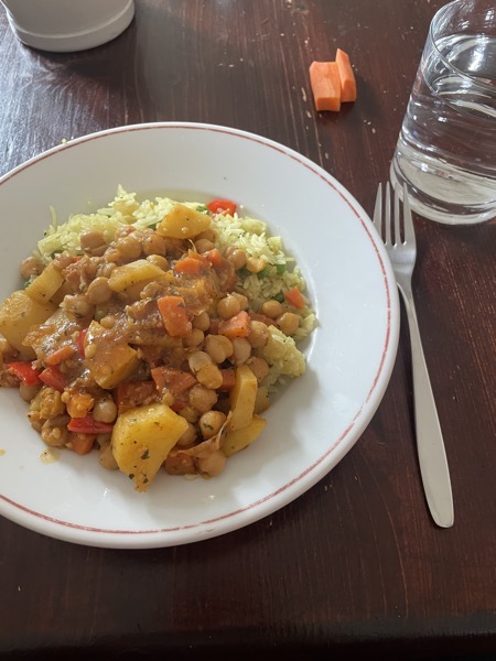 a plate of chickpeas and veggies on top of saffron rice, a glass of water and a few pieces of raw carrot