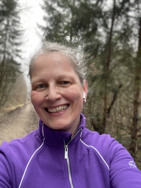 Susanne in a bright purple fleece jacket, her hair pulled back smiling into the camera in front of a trail through the woods