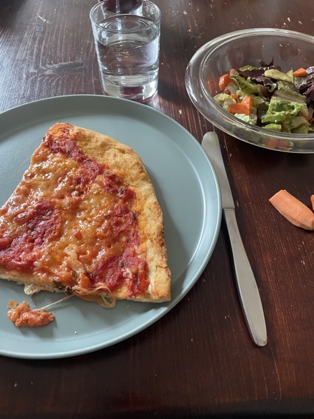 a gray-green plate with a piece of homemade pizza Margherita, a glass of water and a glass bowl of mixed salad on a brown wooden table