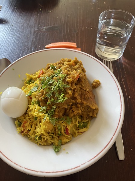A white plate with saffron veggie rice, dal, fresh coriander and a boiled egg, next to it some raw carrot sticks and a glass of water