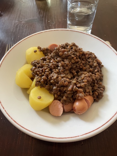 a plate of portatoes, brown lentils and pieces of wiener with a glass of water on the side