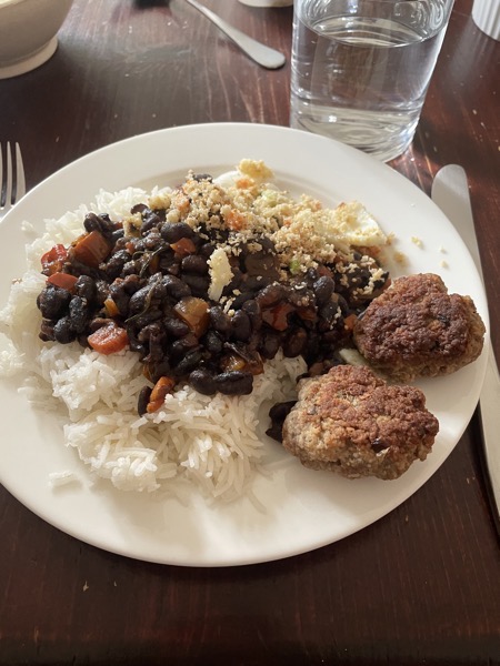 a plate of rice, black beans with veggies, burgers and farofa with a glass of water on the side