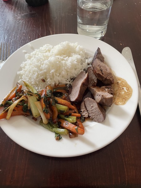 a white plate with rice, julienned veggies, slices of duck breast, sauce and a glass of water on the side