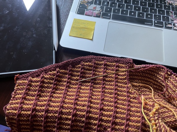 an orange and burgundy half-finished sweater next to an iPad and a laptop