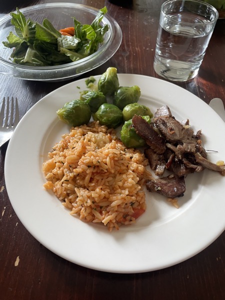 a white plate with kritharaki in tomato sauce, Brussel's sprouts and slices of lamb roast with a bowl of mixed salad and a glass of water
