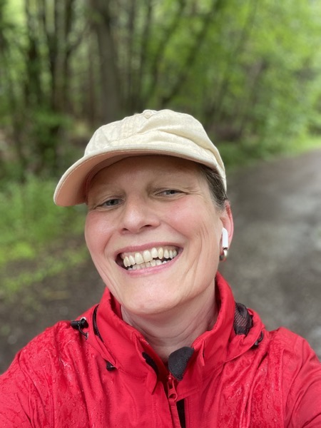a grinning Susanne in a bright red windbreaker and a beige hat in front of some trees everythinng dripping wet